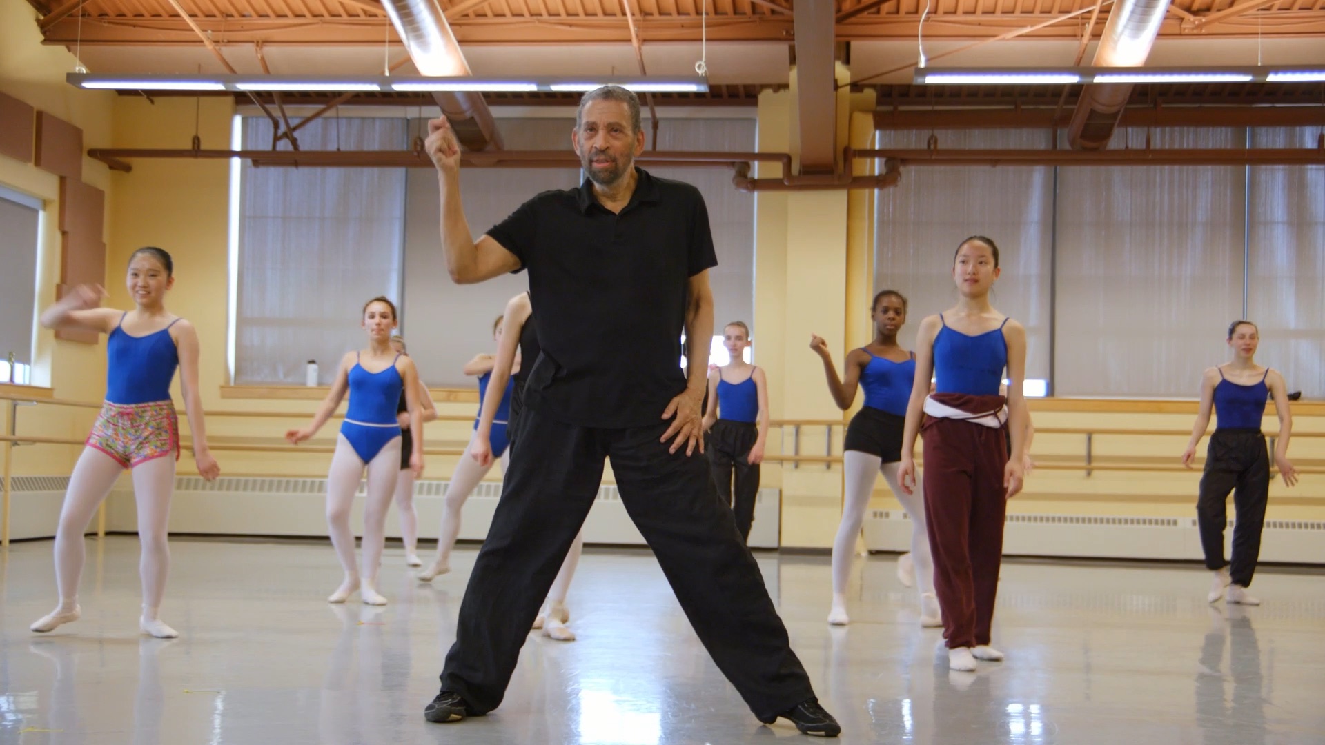 Hines, wearing an all-black outfit and tap shoes, stands at the front of a dance studio full of young students. His legs are in a wide second position, and his right hand is raised, fingers mid-snap.