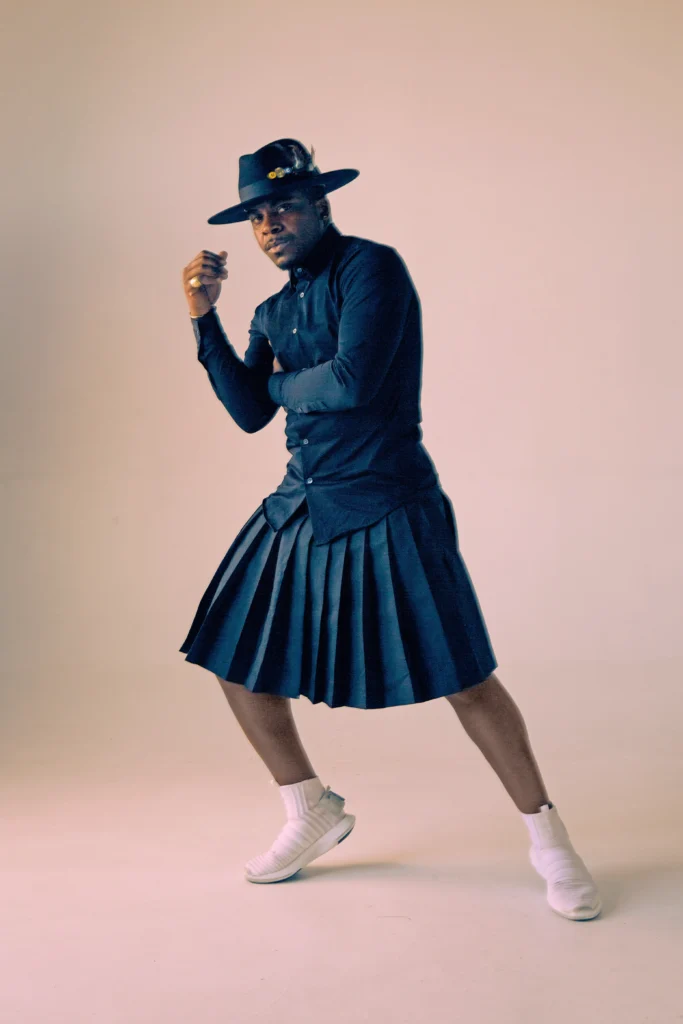 Maleek Washington poses against a pale backdrop. One heel lifts lightly as he slides to the side, an arm crossed over his ribs as the opposite hand rises toward his face. He looks thoughtfully at the camera from under a wide-brimmed hat; He wears a matching dark blue suit with a pleated skirt or kilt and white sneakers.
