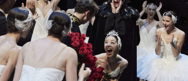 Jill Ōgai, in costume as Odette, bends forward as she clutches a bouquet of roses. She seems to be yelling or laughing in excitement and joy as her castmates surround her cheering and applauding.