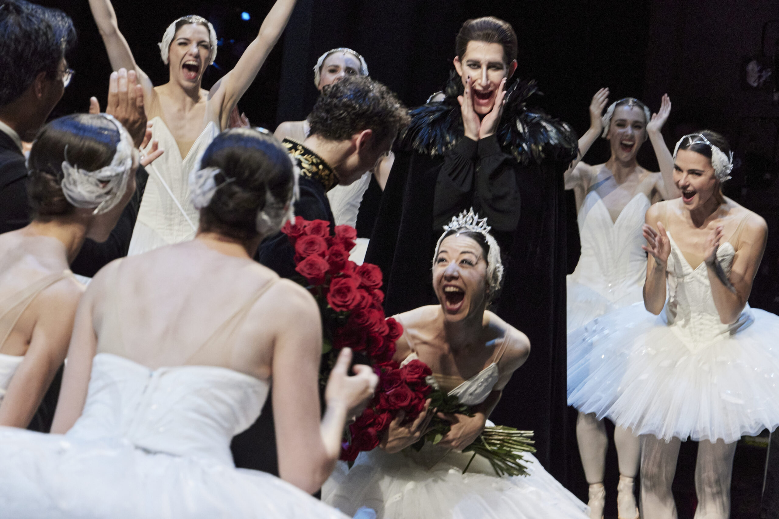 Jill Ōgai, in costume as Odette, bends forward as she clutches a bouquet of roses. She seems to be yelling or laughing in excitement and joy as her castmates surround her cheering and applauding.
