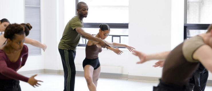 a male instructor guiding a female dancer's arm while taking class in the studio