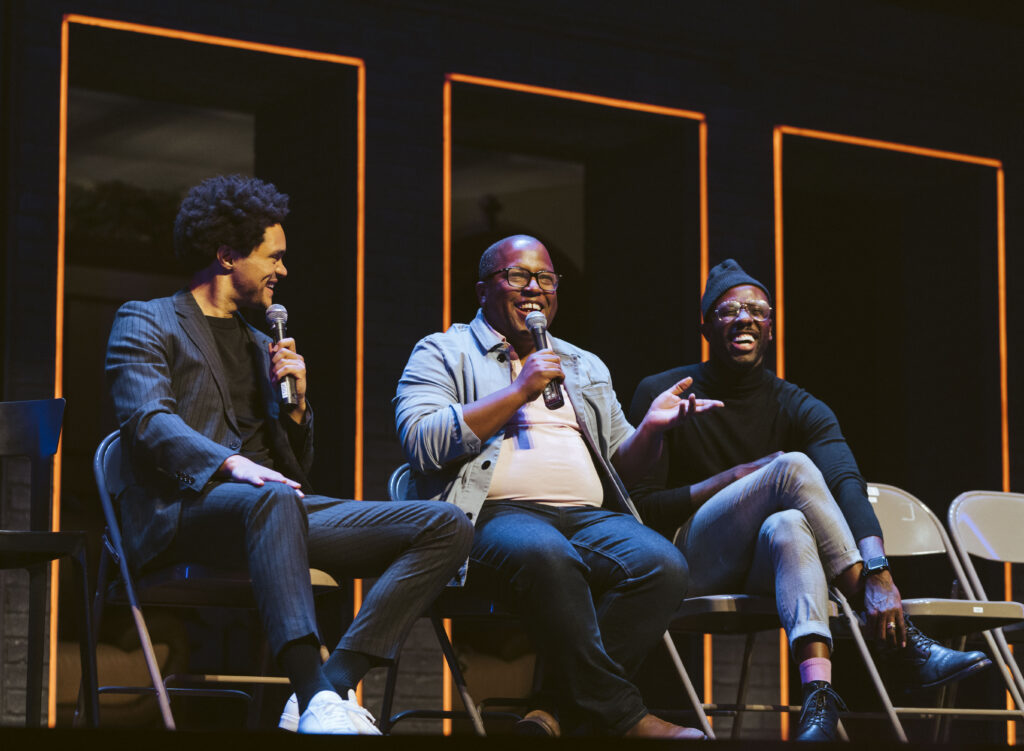 three men sitting in folding chairs on a stage laughing 