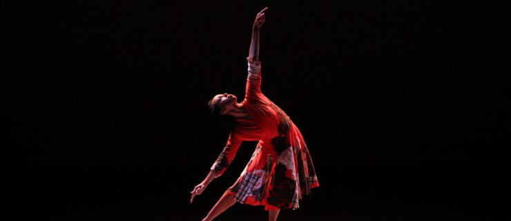 A barefoot dancer caught in a spotlight reaches toward the sky with one arm. She is in plié, one leg extended side so her toes drag along the floor as she bends to the side and reaches. She wears a red dress with large, overlapping patches across the skirt and bodice.