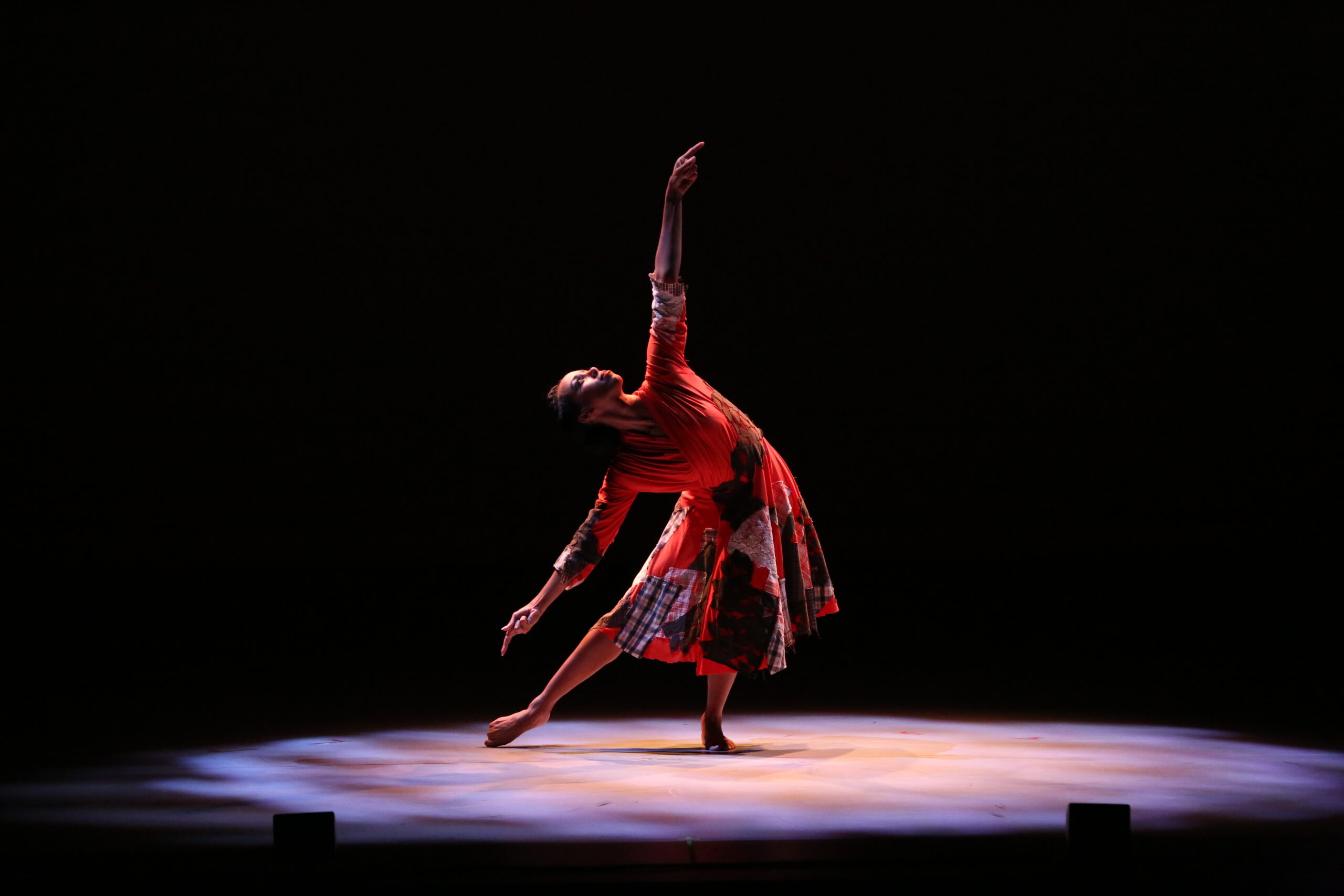 A barefoot dancer caught in a spotlight reaches toward the sky with one arm. She is in plié, one leg extended side so her toes drag along the floor as she bends to the side and reaches. She wears a red dress with large, overlapping patches across the skirt and bodice.