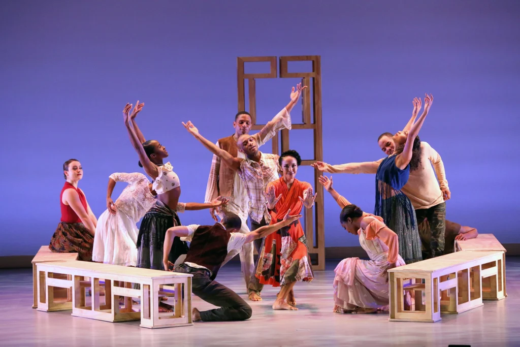 Ten dancers are arrayed on and inside a loose circle of white benches set before a wooden structure upstage. The dancer at the center smiles as she pushes two hands forward, toward the audience. The dancers around her either reach toward her or stretch away.