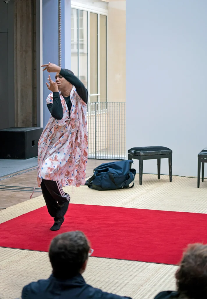 Trajal Harrell performs on a bright red rug. He rises on forced arch one on foot as his knees pull together, arms wrapping up toward his face. He wears a patterned frock over a long sleeved black shirt and black Adidas sweats.