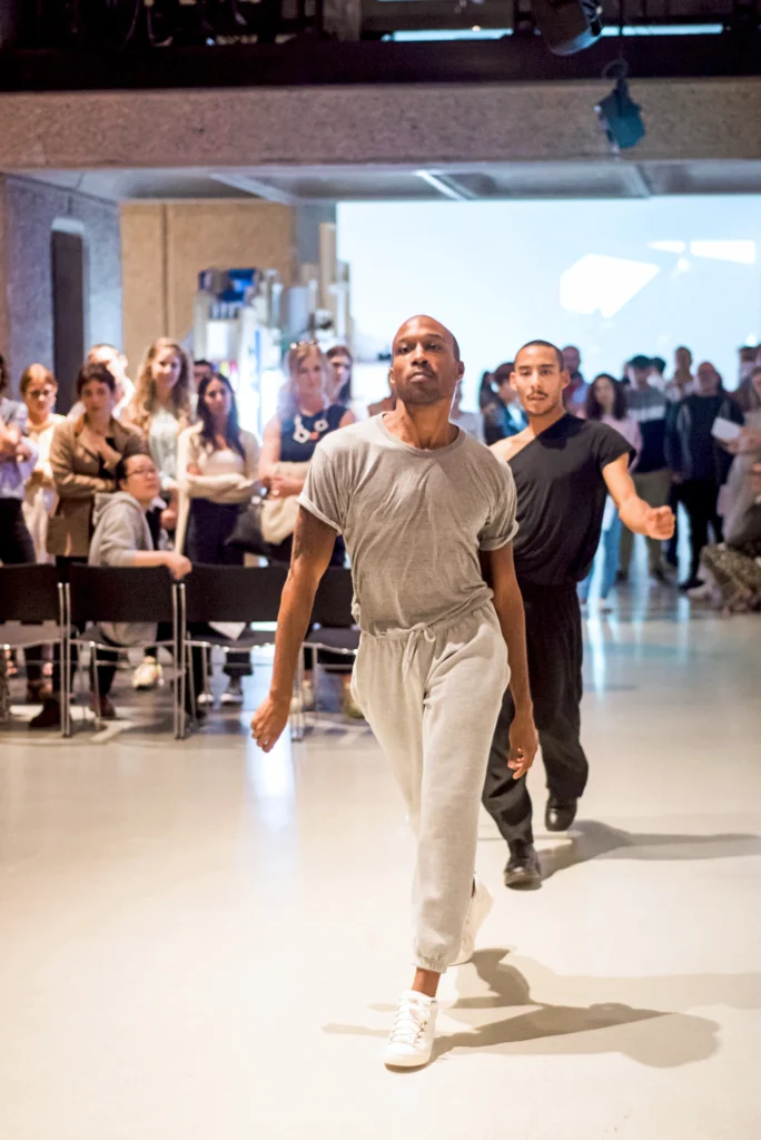 Two dancers walk forward as though on a fashion runway, passing seated audience members who turn to look at them.
