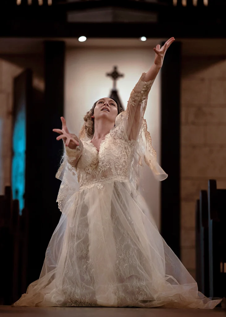 A dancer in an old-fashioned, lacy wedding dress kneels with her arms beseechingly thrust forward, head tipped back as though beseeching something or someone for aid. A blurry cross is visible in the background.