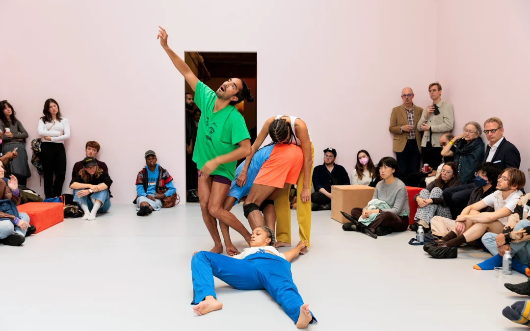 A cluster of four Black dancers costumed in bright colors perform in a white room. One reaches to a corner as their mouth opens in a cry. Another lies on their back, one knee pulled toward their waist. A third bends forward to touch their forehead to another's lower back. Audience members sit and stand around the edges of the space.