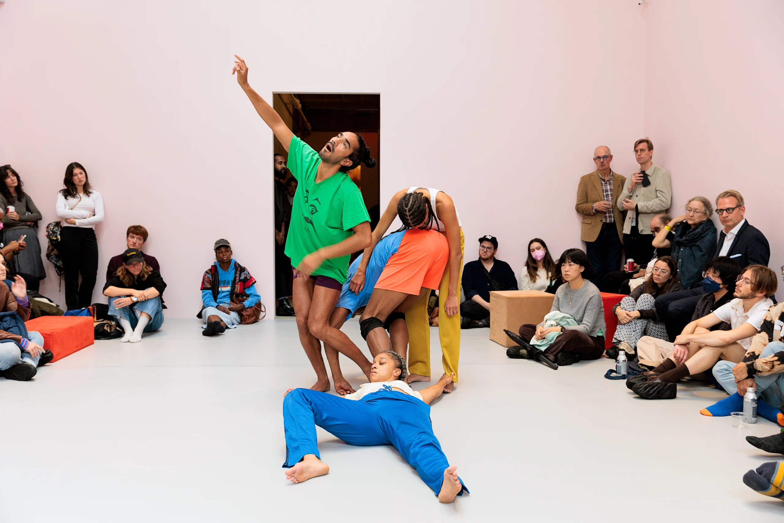 A cluster of four Black dancers costumed in bright colors perform in a white room. One reaches to a corner as their mouth opens in a cry. Another lies on their back, one knee pulled toward their waist. A third bends forward to touch their forehead to another's lower back. Audience members sit and stand around the edges of the space.