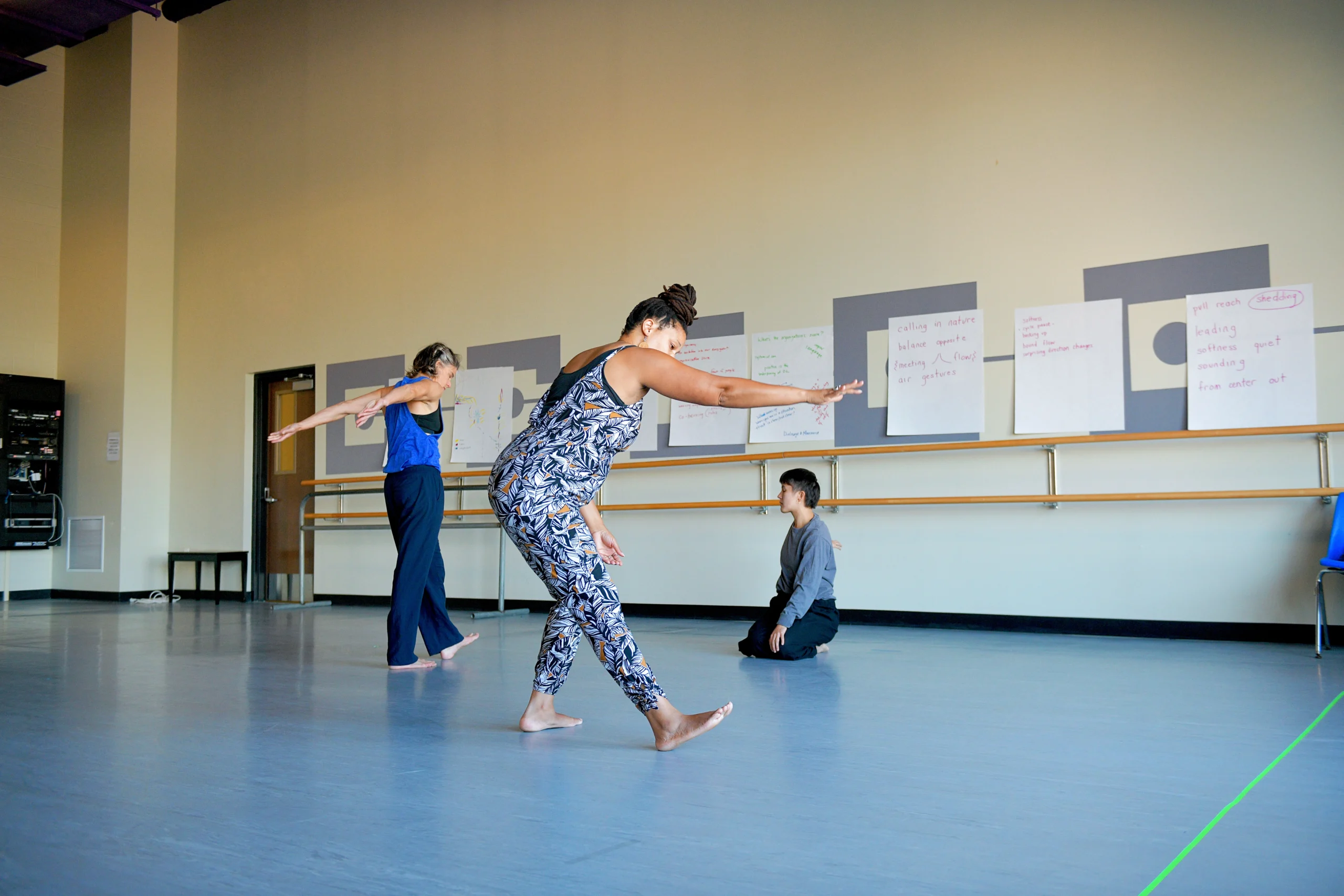 Three dance artists move independently in a studio. One kneels, another swings their arms behind them as they step forward, and a third extends a heel and hand forward, creating straight lines and angles. All are barefoot and wearing warm rehearsal clothes.
