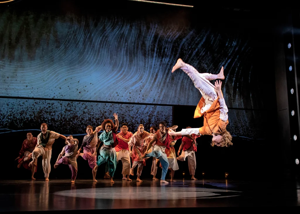 A dancer downstage is captured mid-flip, entirely upside down as he flies through the air. A large group of brightly costume dancers cluster upstage, smiling as one foot raises off the ground in unison.