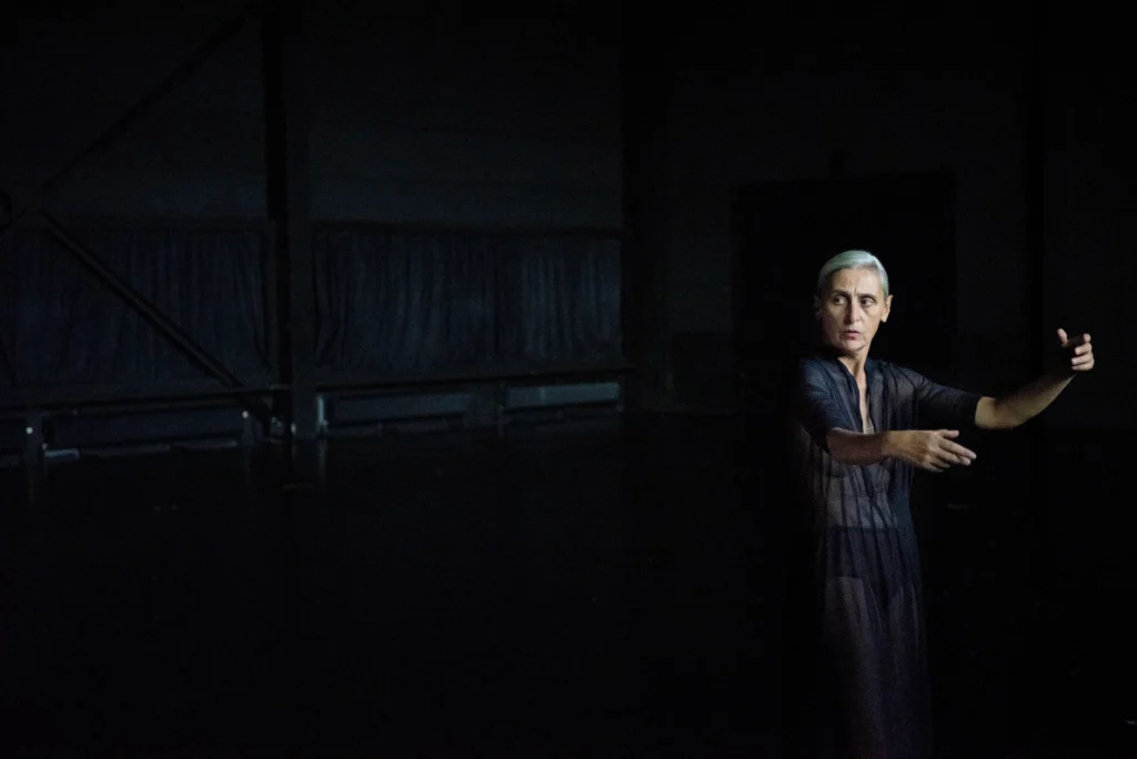 Anne Teresa de Keersmaeker looks over her shoulder on a dark stage. Her arms are softly raised in front of her, torso just beginning to contract. Her grey hair is pulled neatly back from her face; she wears a sheer dark robe over a nude colored tank top and dark briefs.