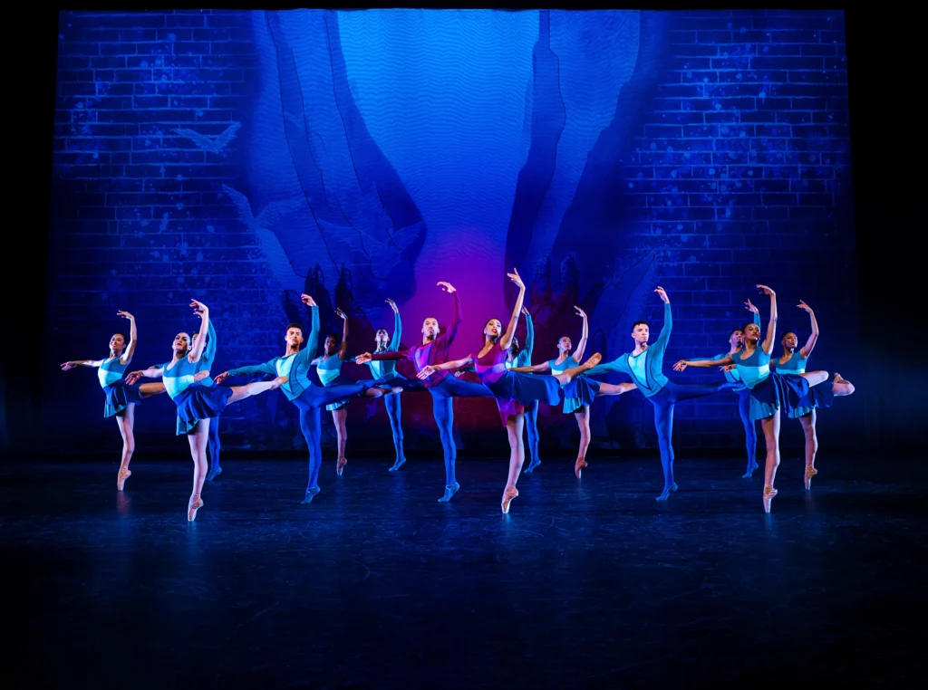 Over a dozen dancers pose in back attitude, the women on pointe, working side arm raised in high fifth. All are dressed in shades of blue, while one male and one female dancer near center have purple tops.