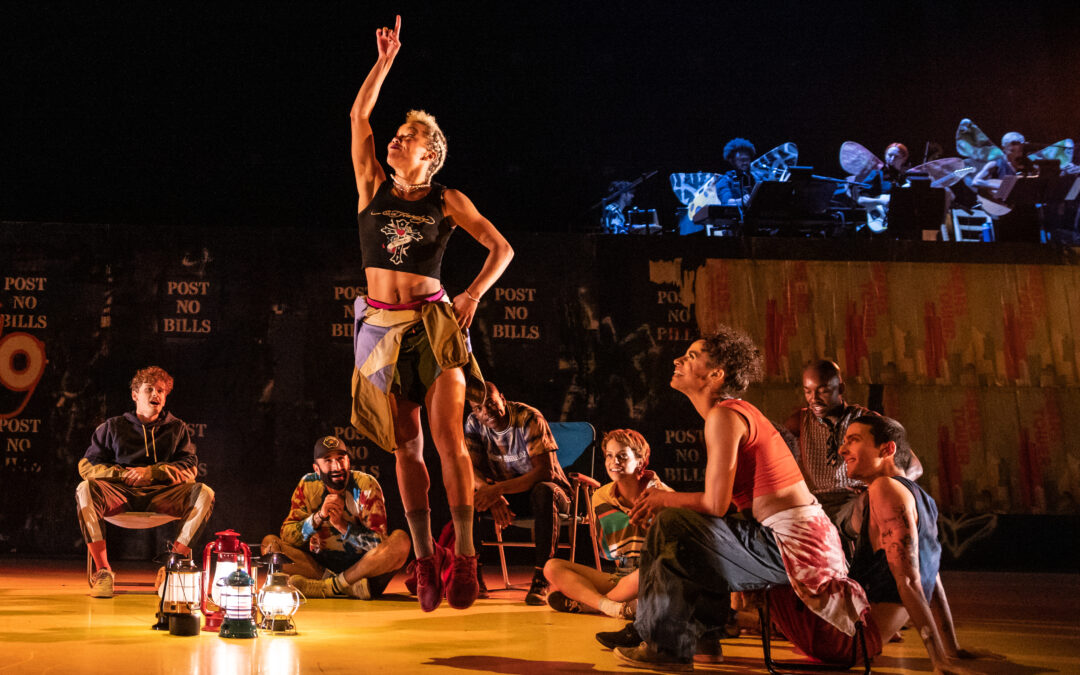 A group of dancers sits in a semicircle around an onstage campfire, grinning at a dancer in the center of the group who is jumping joyously and pointing to the sky.