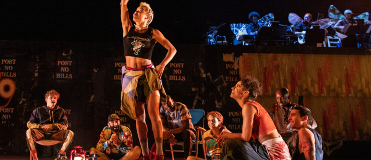 A group of dancers sits in a semicircle around an onstage campfire, grinning at a dancer in the center of the group who is jumping joyously and pointing to the sky.