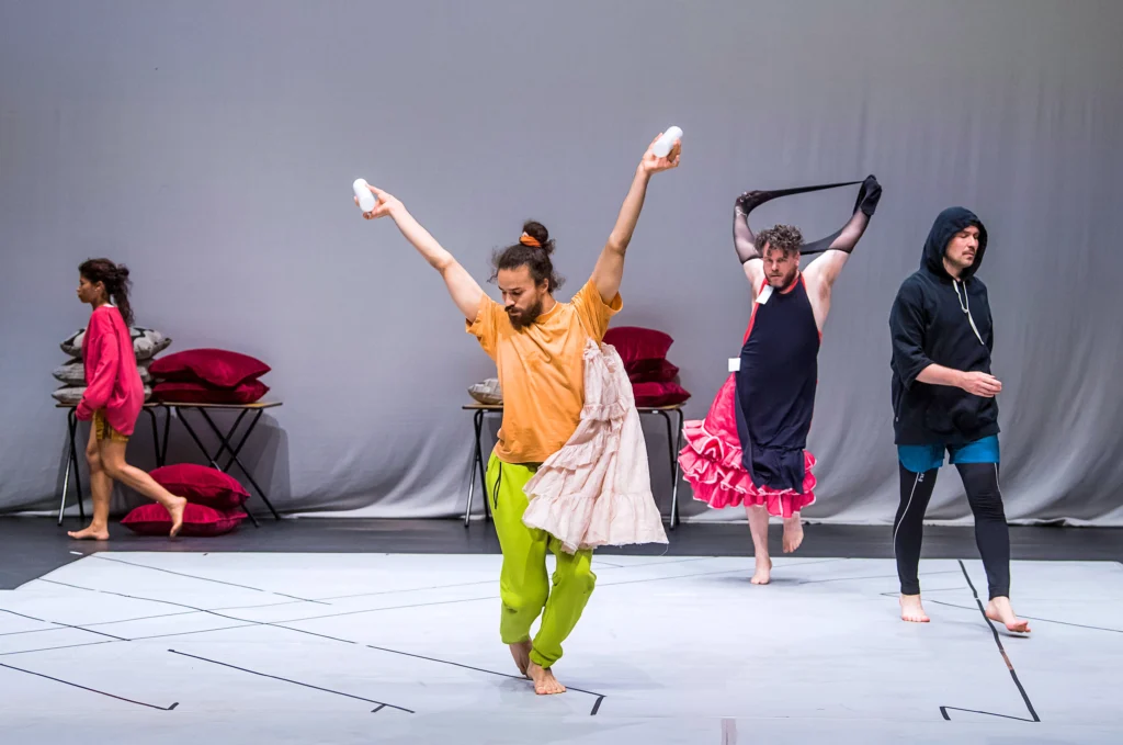 A dancer in a peach t-shirt and bright green sweats poses center stage with arms in a V overhead, holding a pair of white cylinders. Another dancer upstage has black tights tangled around their arms. Two other dancers in rehearsal wear walk around the edges of the space. Tables and chairs are piled with red velvet pillows upstage.