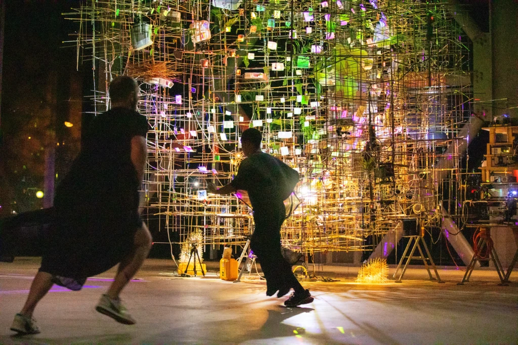 Two dancers blur as they run in the space in front of a wire cage or matrix, to which numerous small rectangles are affixed. The structure is warmly lit in yellows, purples, and green.