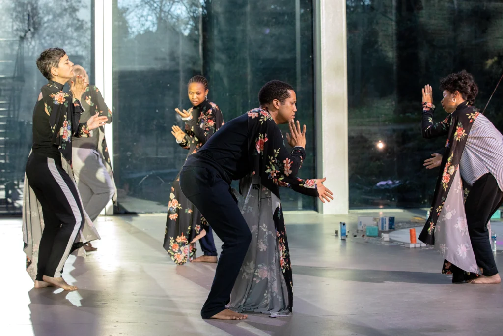 A loose circle of five dancers bend forward as they clap in unison, facing different directions. All are draped in black fabric with reddish pink flowers.