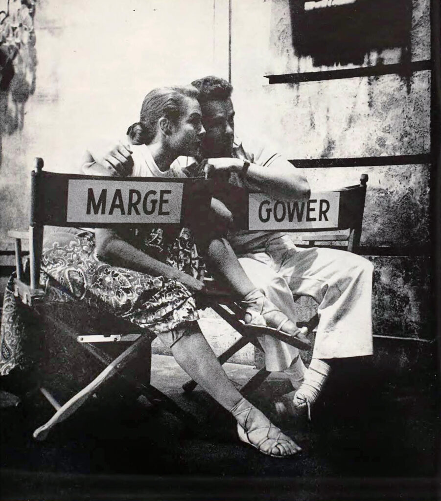 In a black and white archival photo, Marge and Gower sit backwards on directors chairs emblazoned with their first names. His arm is around her shoulders as they put their heads together, both studying something off camera. Marge has one knee pulled up to her chest; their legs almost seem to tangle.
