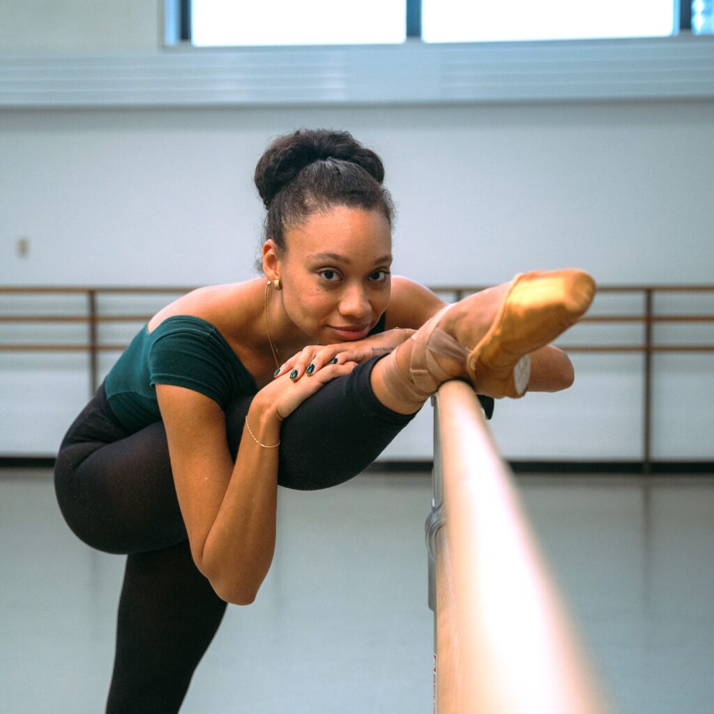 a ballerina stretching at the barre with her leg extended forward and resting her hands and chin on her leg