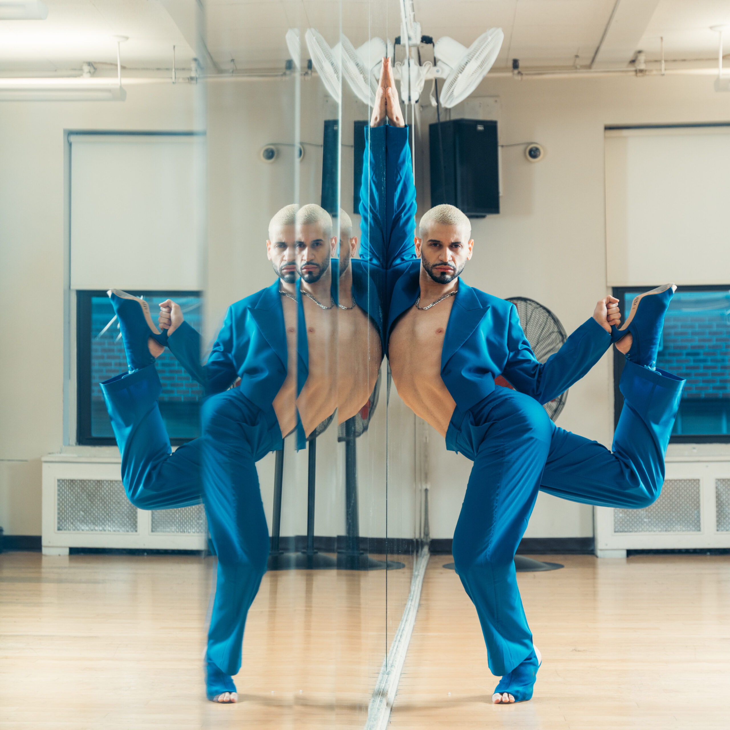 a male dancer wearing a blue suit and heels posing against a mirror in a studio