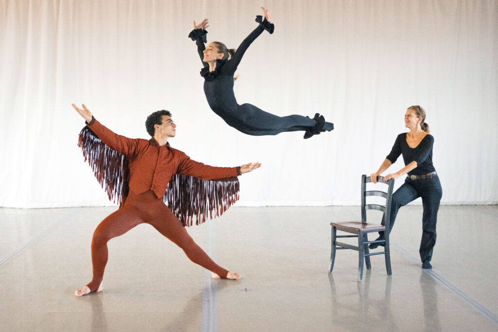 a male dancer wearing a reg costume with wings about to catch a female dancer in all black jumping off a chair that is being held steady by another woman