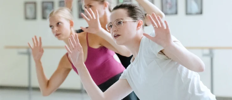 Emma Portner, in a white t-shirt and glasses, is shown from the waist-up. She leans to one side, hands raised as though pressing them against glass. Two dancers just behind her imitate the pose as they stand back-to-front, very close together.