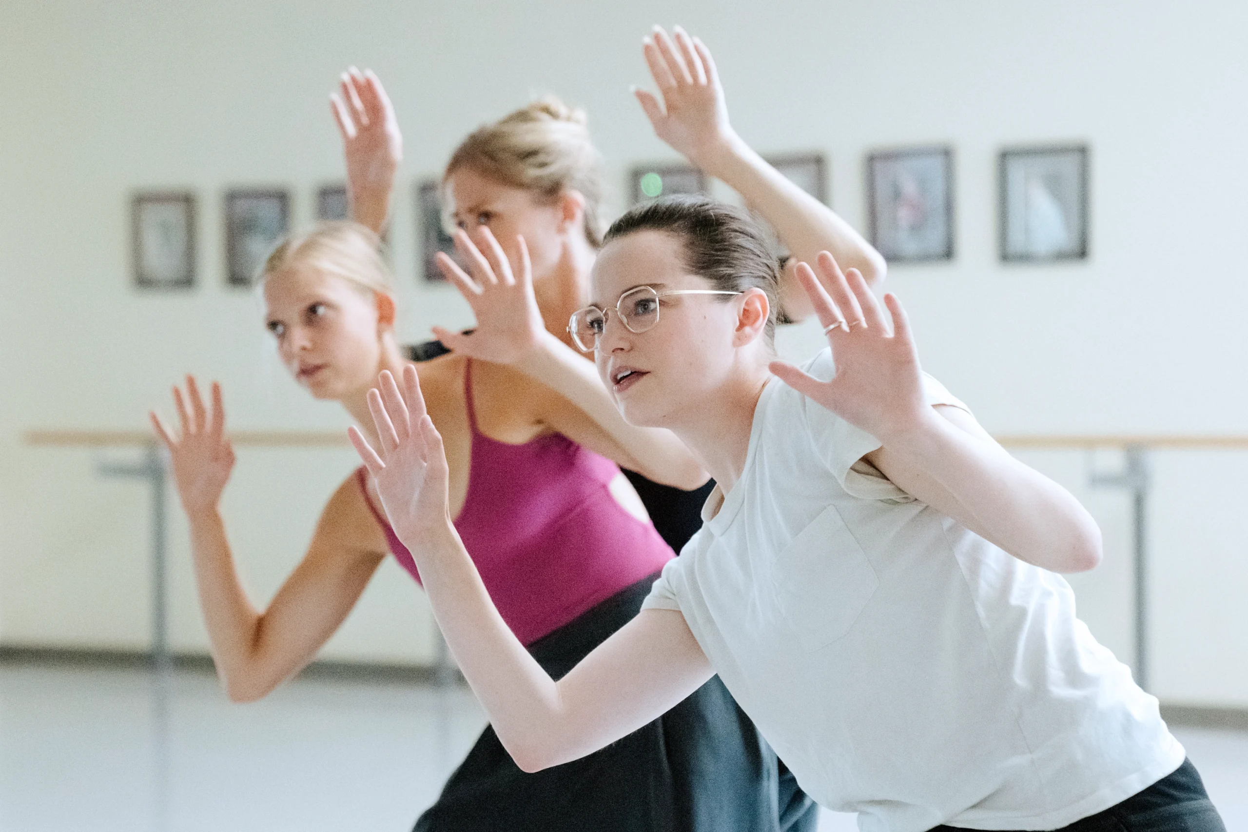 Emma Portner, in a white t-shirt and glasses, is shown from the waist-up. She leans to one side, hands raised as though pressing them against glass. Two dancers just behind her imitate the pose as they stand back-to-front, very close together.