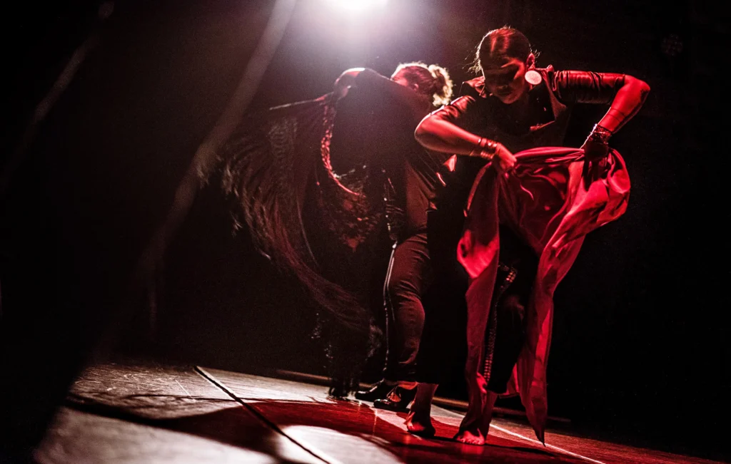 Arielle Rosales and Brinda Guha are a blur of motion in red light. Rosales whirls a tasseled cape before her, while Guha uses both hands to lift her skirts, gazing over at her bare feet.