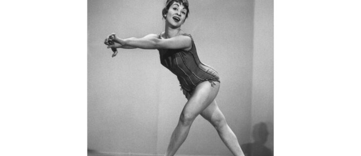 A black-and-white photo of Chita Rivera. Wearing a seamed leotard and pumps, she crosses her right leg behind her left and extends her arms to her right, her mouth slightly open, as if she's mid-song.