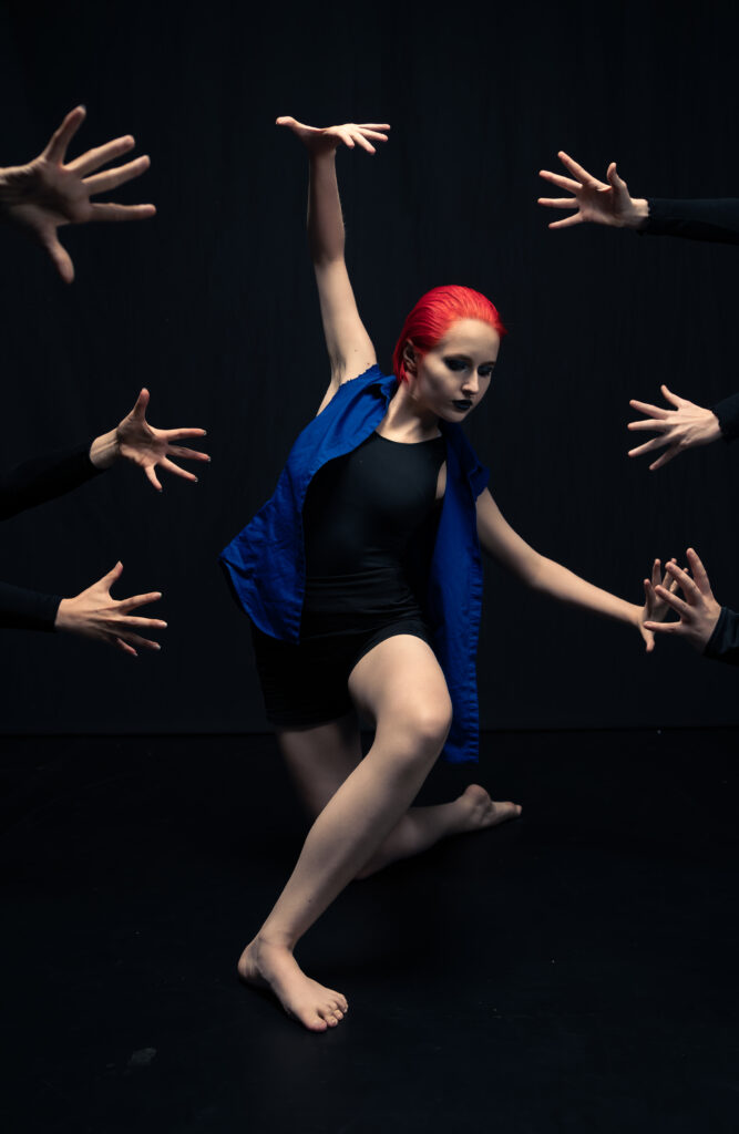 a female dancer with bright red hair posing with her arms out while many heads reach towards her