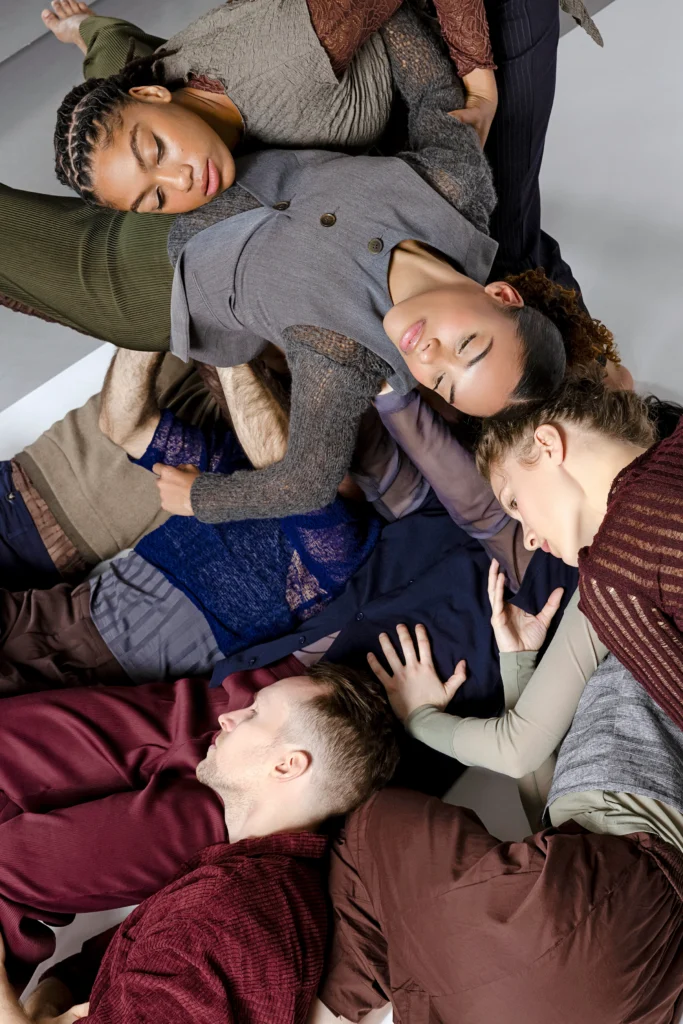 Seven dancers pile and curl atop each other on the floor, heads resting on chests and hips. They wear knits and layers in shades of reds, greys, and blues.