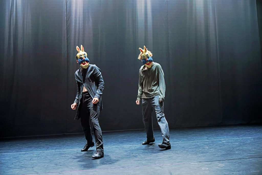 Two dancers in mildly scary bunny masks rock onto their back feet as they stare forward.