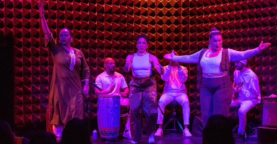 Brinda Guha, Amanda Castro, and Arielle Rosales are lit in pink and purple light as they perform as Soles of Duende at the front of a small stage, two seated percussionists and a guitarist visible behind them. Guha raises an arm to the sky as she looks intensely out at the audience. Castro looks focused as she taps, hands separated but poised to clap. Rosales opens her arms wide in invitation, chin ducked as she looks challengingly out at the audience.