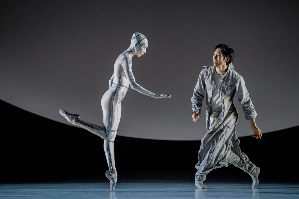 A ballerina in a silver jumpsuit balances en pointe; she appears to be an android. A male dancer watches her with a look of fascination and excitement as he moves toward her.