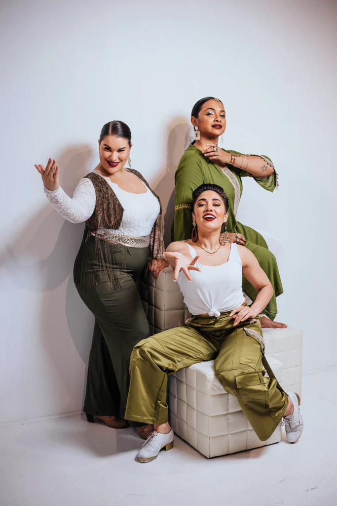 Arielle Rosales, Brinda Guha, and Amanda Castro (Soles of Duende) pose together, all wearing shades of green and white. Rosales smiles cheekily, chin ducked and an arm elegantly curved, palm up in invitation. Castro, seated, lifts her chin and smiles brightly, one hand outstretched palm up to the camera, knees bending as though ready to begin tapping any second. Guha sits elevated behind them both in profile, an inviting smile on her face as she gracefully crosses one arm to touch the opposite shoulder.