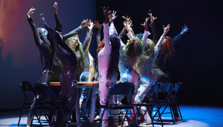 a group of dancers surrounding a table standing out of their chairs with arms lifted overhead