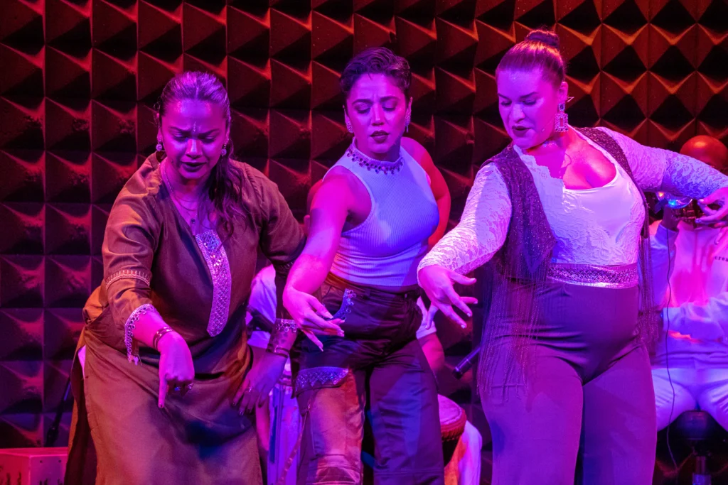 Brinda Guha, Amanda Castro, and Arielle Rosales stand close together in a Soles of Duende performance. Each extends their right arm forward to the center of their front-facing cluster, fingers closing in a manner familiar to flamenco technique. They are lit in purples and pinks on a small stage with a textured, dark back wall.