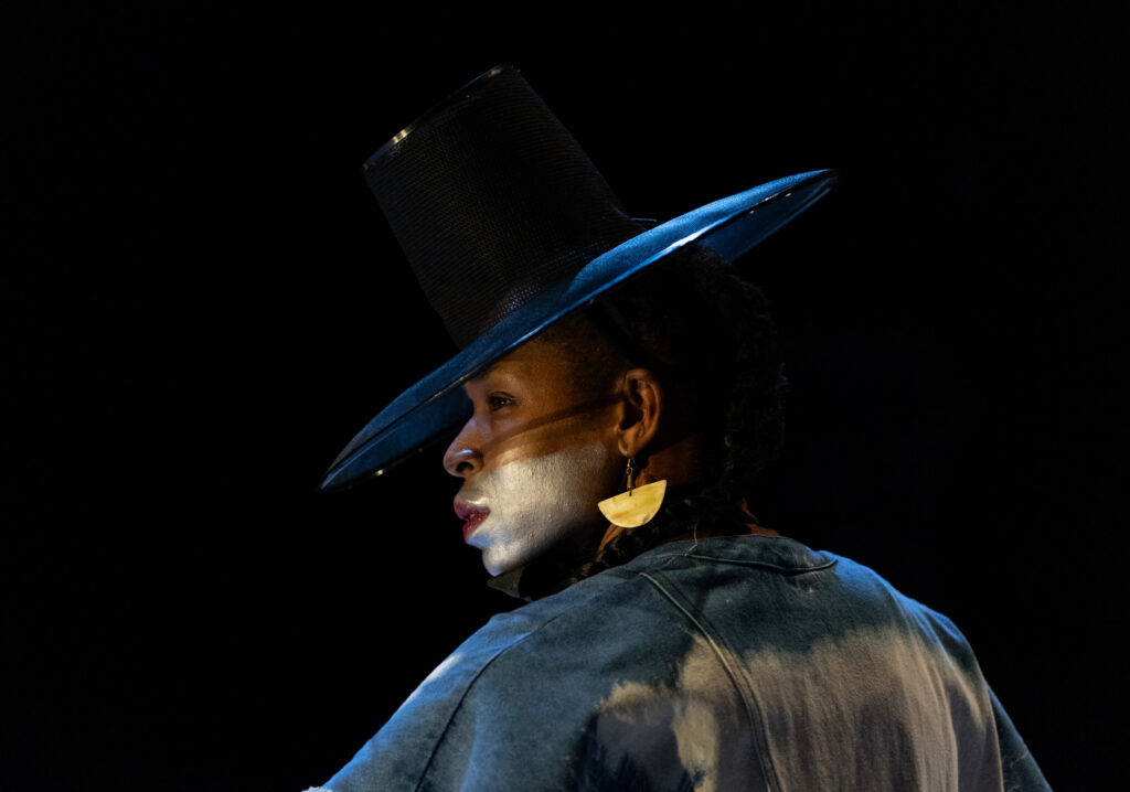 Marjani Forté-Saunders is photographed in profile. Her back is turned to the camera and she looks to the left from beneath a wide-brimmed hat, eyes intent. The bottom half of her face is painted silver; a yellow half moon earring dangles from her visible ear.
