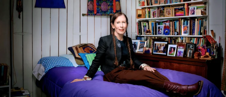 Meredith Monk sits on a bed covered in a purple duvet. Here reddish brown hair is in twin braids resting on either shoulder. She wears a black jacket and red-brown trousers and boots. To one side is a set of shelves teeming with colorful books and framed photos. On the walls are tapestries and images of Hindu deities. The walls are white and slatted.