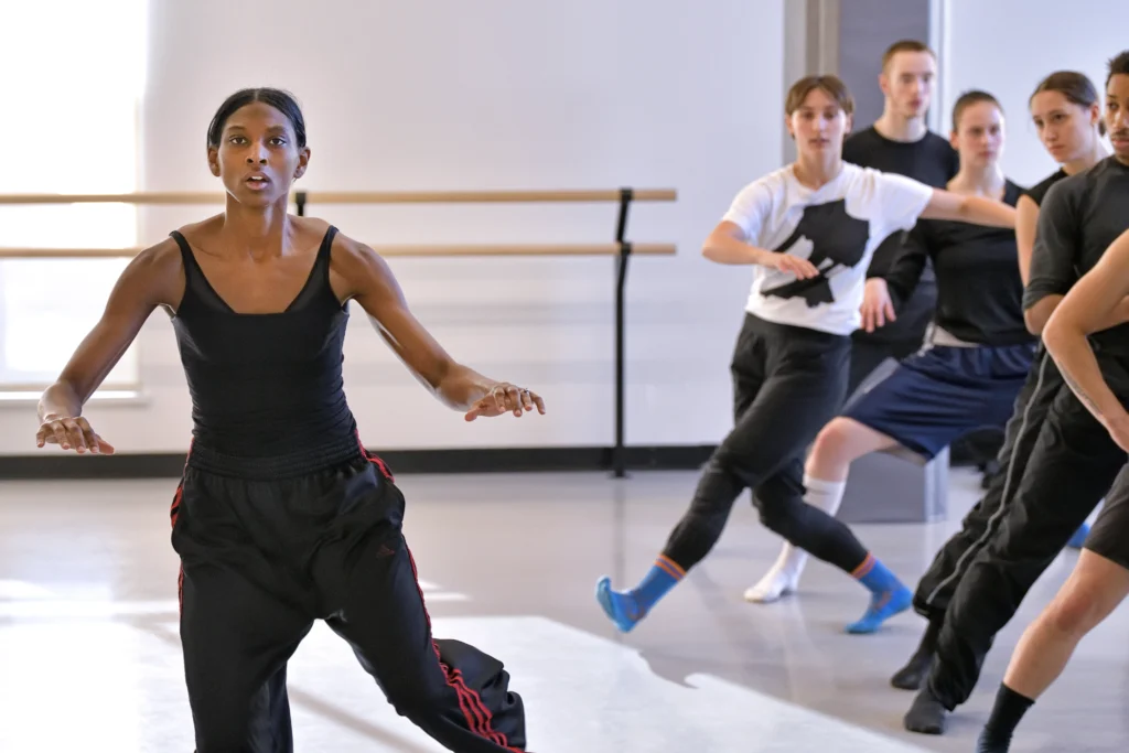 Micaela Taylor is intensely focused as she rests her hands at hip height, moving onto her right foot. To her left, a half dozen dancers in rehearsal gear imitate her movement in a vertical line.