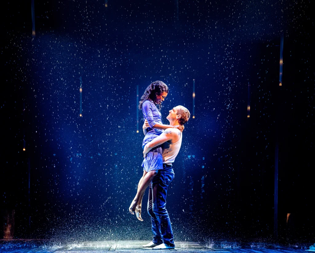 A man in jeans holds a barefoot woman in a dress up, his arms curved around her hips and waist. They smile at each other as rain splashes around them.