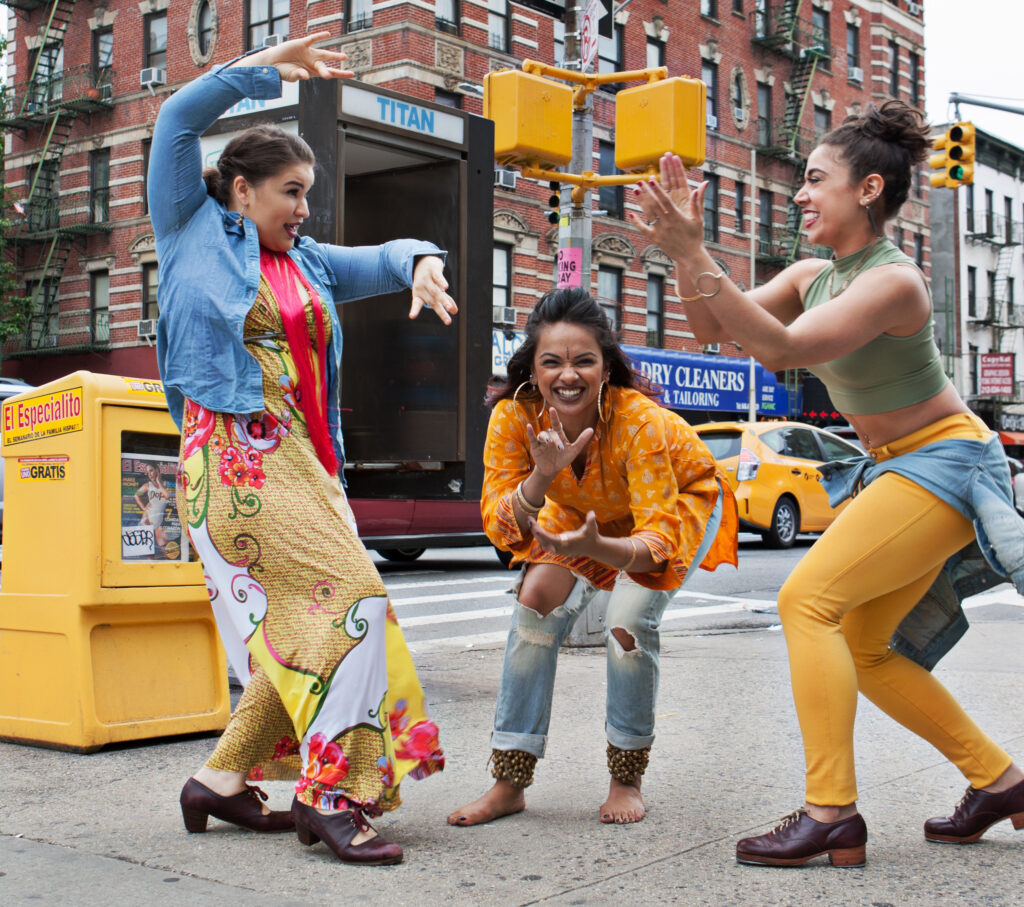 Arielle Rosales, Brinda Guha, and Amanda Castro (Soles of Duende) jam together on a New York City street corner. Guha, in center, leans forward and grins at the camera, nose scrunching, as she claps; she is barefoot, and wears ghungroo ankle bells. On either side, Rosales and Castro face each other, Castro grinning as she claps and stamps in her tap shoes, Rosales giving a playful look as she raises her arms overhead, flamenco shoes ready to drop a heel.