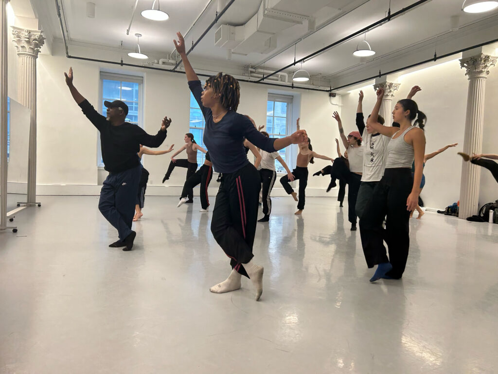 a group of dancers in a white walled studio learning choreography 