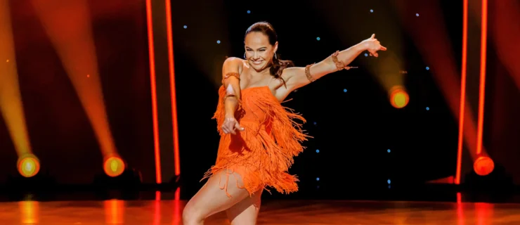 Alexis Warr performs solo on the "So You Think You Can Dance" soundstage. She wears heels and a short orange dress that moves with her.