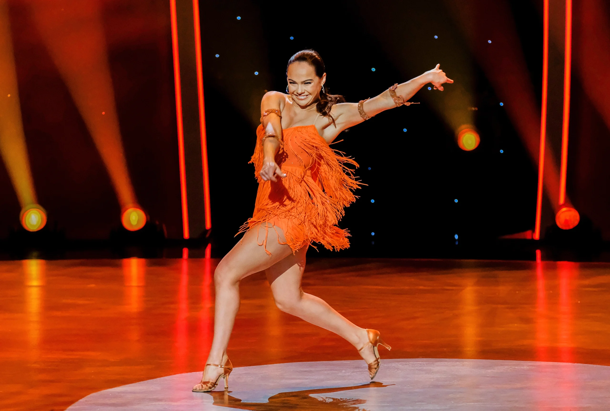Alexis Warr performs solo on the "So You Think You Can Dance" soundstage. She wears heels and a short orange dress that moves with her.