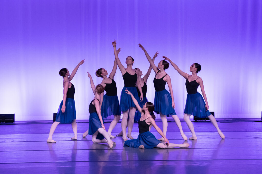 a group of female dancers wearing black leotards, blue ballet skirts, pink tights and shoes, posing on stage with a purple backdrop 