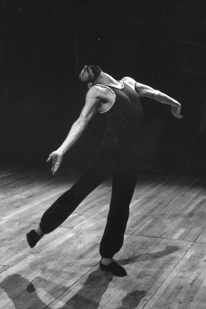 A black and white photo of Paxton, wearing a dark unitard, in an ecstatic low arabesque, his palms raised as if in supplication, his head thrown back.