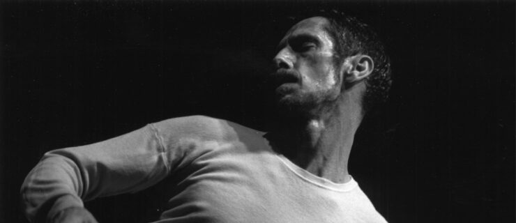 A black and white photo of Paxton from the torso up. Wearing a light-colored long-sleeve t-shirt, he twists his body to the right, his eyes looking down and out over his right shoulder, his brow furrowed in concentration.